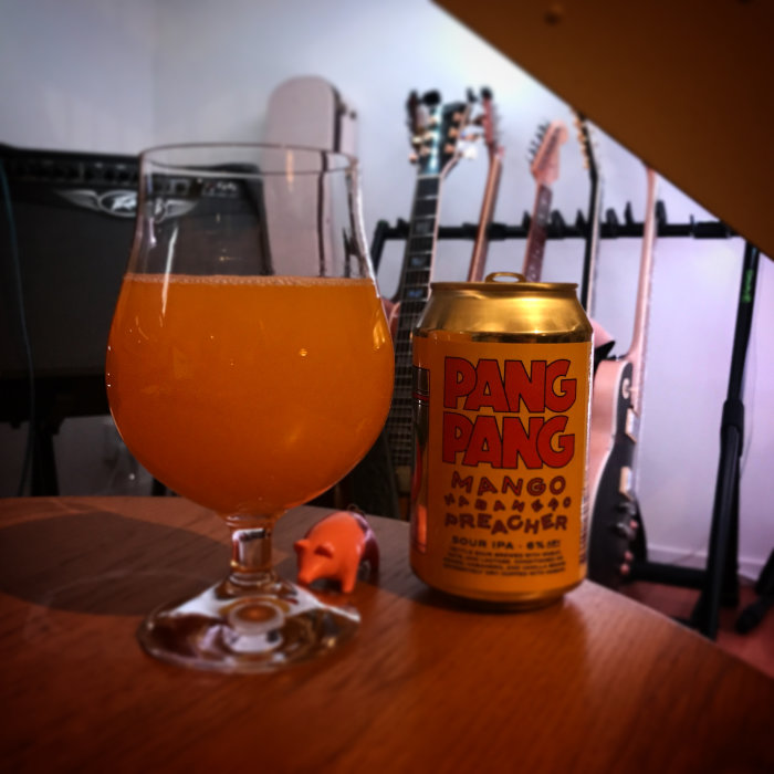 Ett glas med orange dryck bredvid en ölburk med texten "PANG PANG MANGO PREACHER" och gitarrer i bakgrunden.