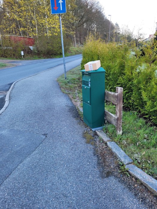 Paket levererat ovanpå postlåda vid trottoarkant med förbipasserande väg och växtlighet.