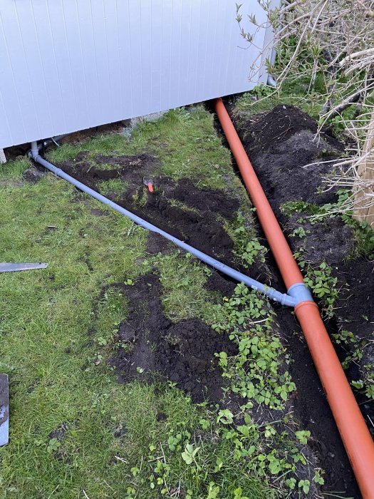 En grå och en röd avloppsrör anslutna med lutning i en grävd ränna vid en byggnads kant.