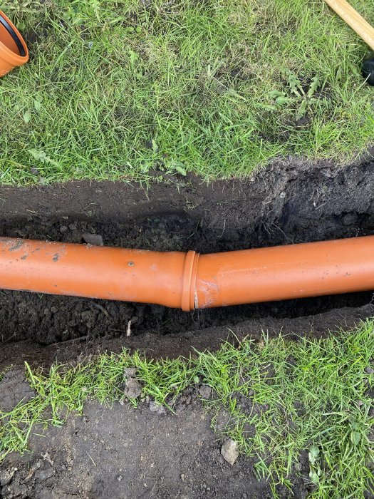 Orange dräneringsrör som sammanfogats under en vinkel, nedgrävda i en grävd ränna, omgivna av gräs.