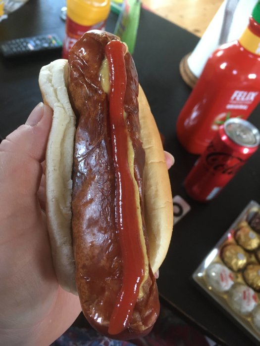 En grillad korv med bröd och ketchup i en hand, med matprodukter i bakgrunden.