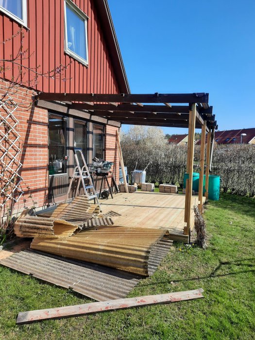 Delvis rivet träntak med staplar av gammalt och nytt virke framför en ny pergola intill ett rött hus.