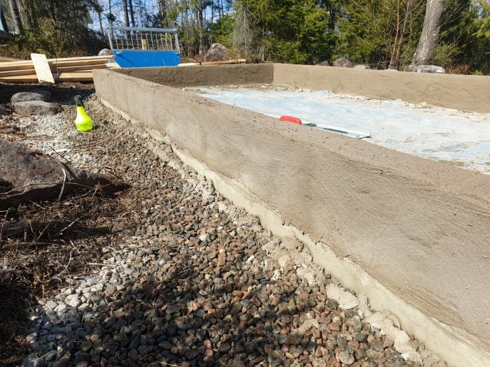 Nypustad och oregelbunden betongvägg med verktyg och brädor i en byggmiljö under soligt väder.