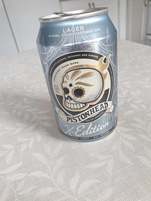 Ölburk med dödskallemotiv, märkt "Pistonhead Lager", placerad på ett bord med mönstrad duk.