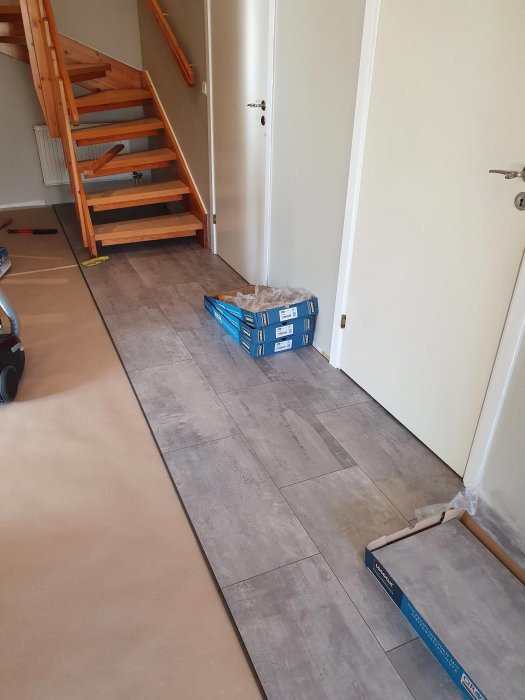 Nytt laminatgolv läggs i en hall med en öppen trätrappa och en låda kakel på golvet.