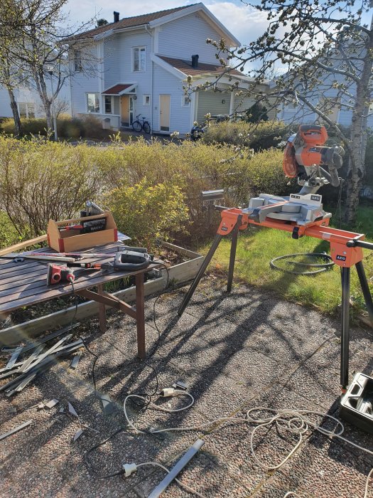 Arbetsområde utomhus med såg på stativ, verktyg och material framför ett vitt hus i soligt väder.