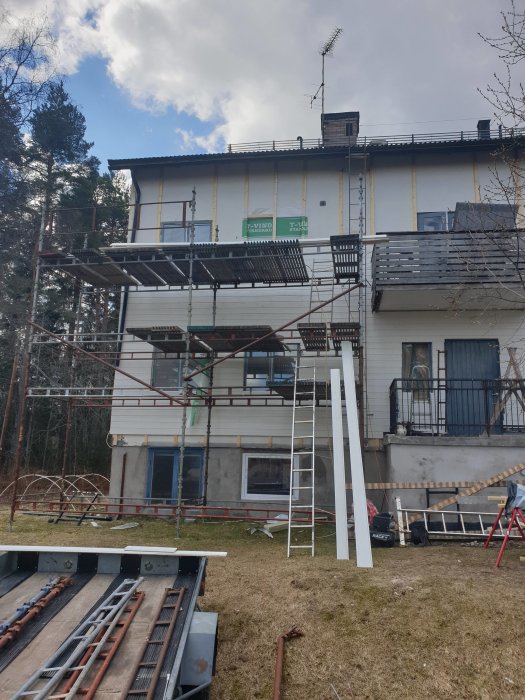 Ställning uppställd vid vit husfasad under renovering med olika byggmaterial i förgrunden.