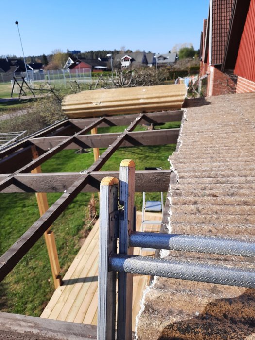 Rivet altantak med synliga träbjälkar och stålbalkar på en bakgård med grönt gräs och hus i bakgrunden.