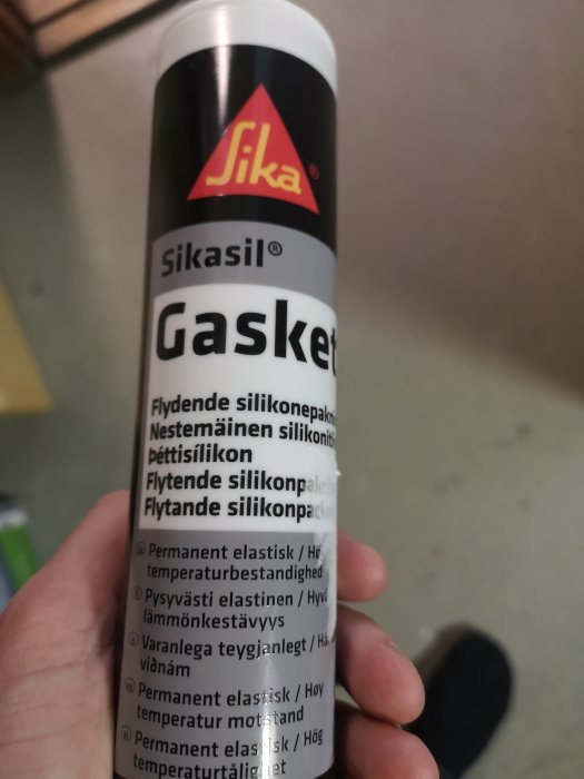 Hand håller en flaska Sikasil Gasket silikonpackning mot en suddig bakgrund.