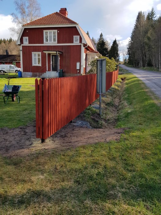 Nybyggt rött staket längs gräsmatta med hus och träd i bakgrunden.