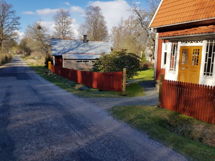 Nyuppsatt rött trästaket vid sidan av en grusväg bredvid ett traditionellt rött hus med vita knutar och gult dörrparti.