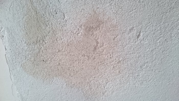 Grov, ujævn væg med huller og striber fra pudsning, set i sollys; pletvis misfarvning og ujævn tekstur tydelig.