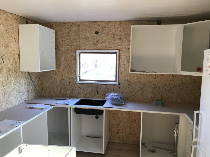 Kök under renovering med osb-väggar, ett mindre fönster och vita skåps stommar utan dörrar.