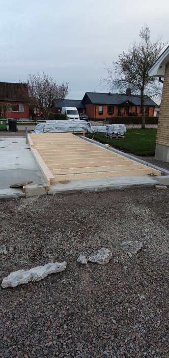 Påbörjad trästomme för byggprojekt med uppmätta och kapade reglar på en betonggrund, omgiven av grus och bostadshus.