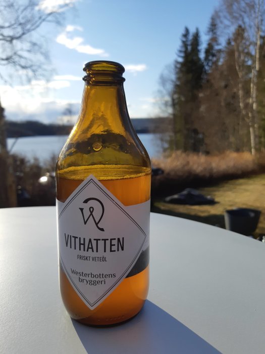 En brun glasflaska märkt "VITHATTEN FRISKT VETEÖL" på ett bord utomhus med sjö och träd i bakgrunden.