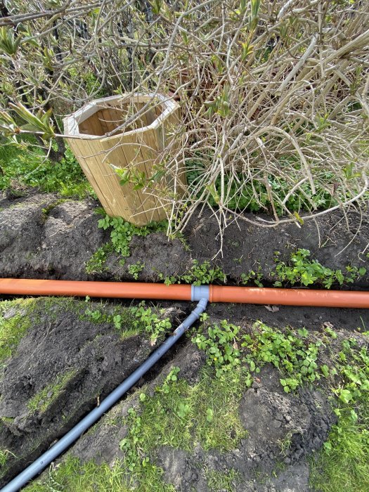 Orange och blå rör anslutna med kopplingar i en grävd ränna i trädgården.