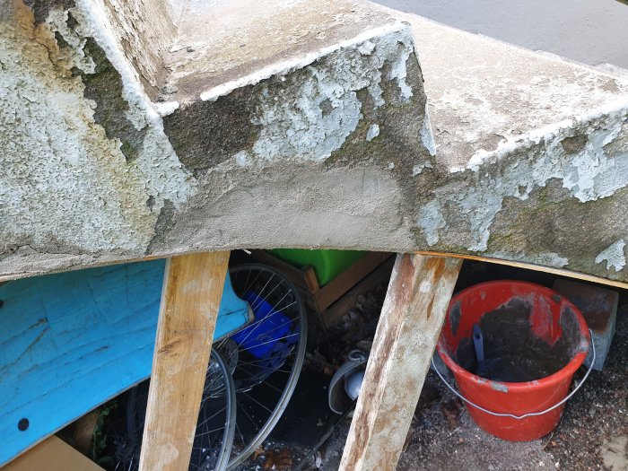 Slidt cementtrappe med afskallet maling, understøttet af træplanker. Rød spand med byggematerialer og en cykel under trappen.