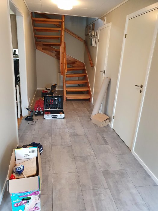 Renoverad hall med grått klinkergolv och trätrappa, lister och kartonger på golvet.