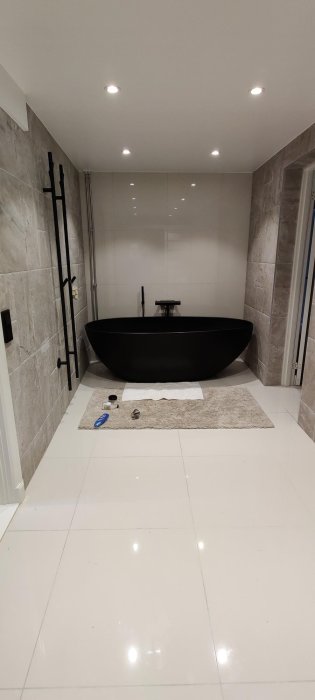 Modernt badrum med fristående svart badkar, kaklade väggar och infälld belysning.