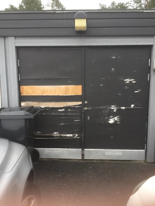 Nött svartmålad garagedörr med synlig sliten faner och en plywood-lapp.