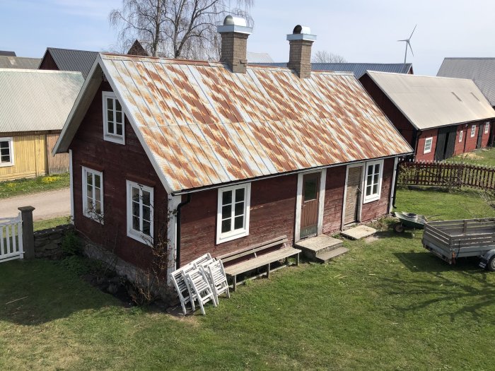 Röd stuga med slitet zinkplåttak, omgiven av andra byggnader och en vindkraftverk i bakgrunden.