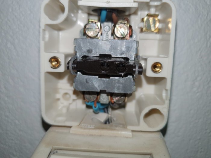 Öppen väggströmbrytare med synlig kabelanslutning och mekanism, bakom vit plastdosa.