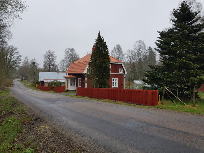 Nytt rött staket längs vägkanten framför en röd husfasad under en regnig himmel.