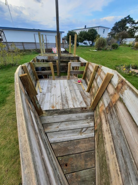 Lekbåt under konstruktion i trä, uppbyggd i en trädgård, med bostadshus i bakgrunden.