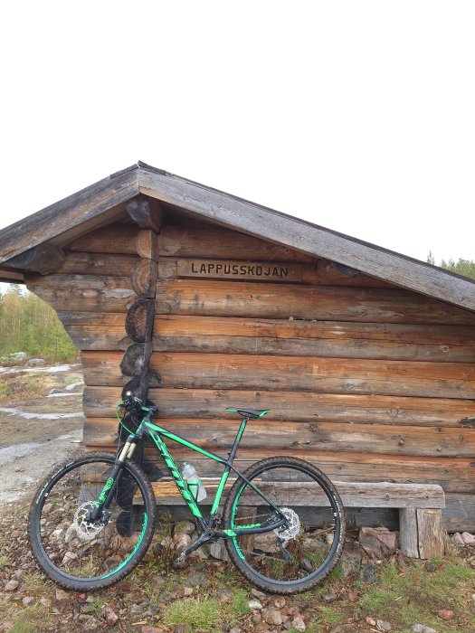 En grön mountainbike lutad mot en rustik timrad stuga med namnet "LAPPUSSKOJAN" i skogsmiljö.