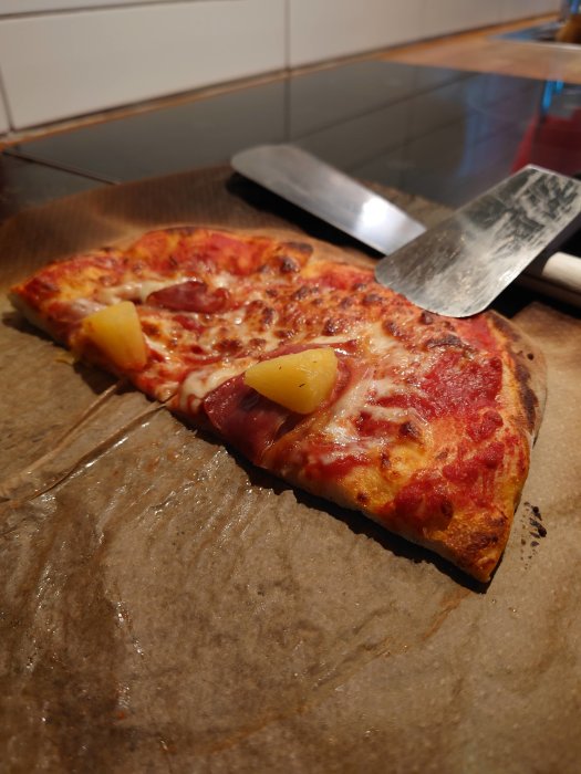 Två skivor pizza med skinka och ananasbitar på bakplåtspapper, pizzaspaden i bakgrunden.