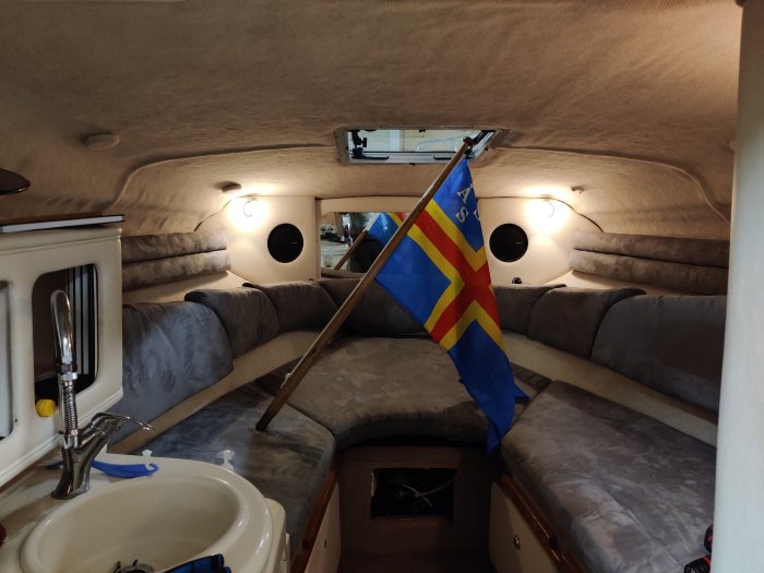 Interiör av båtkabin med Åländsk flagga, sittplatser, ljusarmaturer och ett litet kök.