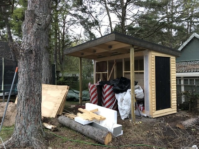Nybyggt trähus med tak målat med tjärvitrol, byggmaterial framför och träd i bakgrunden.