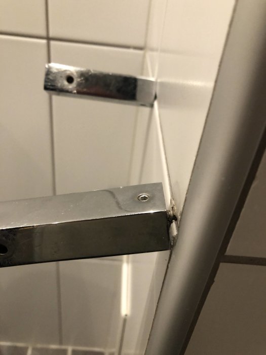 Toalettpappershållare av metall som delvis lossnat från vit kakelvägg.