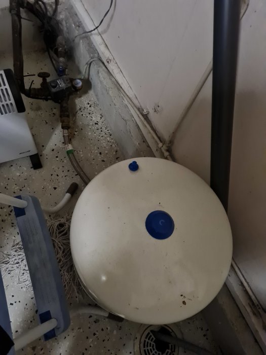 En vit hydropress behållare och diverse vattenledningar och anordningar på ett smutsigt golv.