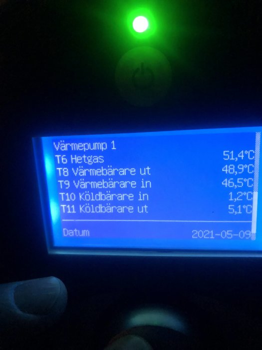 Display av värmepump med temperaturdata och datum, grönt ljus och på-knapp i mörker.