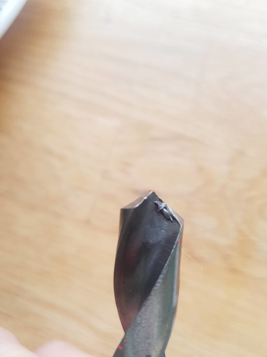 Närbild på en defekt borrspets med synliga skador och dålig slipning, mot en oskarp träbakgrund.