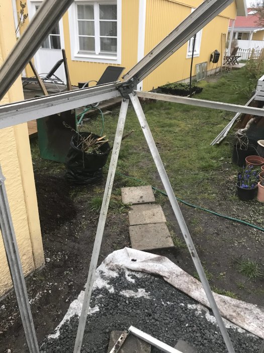 Delvis uppbyggt växthus med ovanliga hörnbeslag i en trädgård, grå stomme utan glas, mot en gul husvägg.