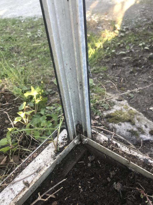 Närbild på hörnet av ett äldre växthus med distinkta metallhörnbeslag, omgiven av jord och vegetation.