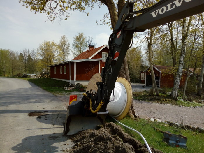 Grävmaskin från Volvo gräver för fiberinstallation vid röd villa med vita knutar.