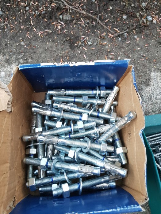 En kartong full med nya expanderbultar av metall på en grusig mark.