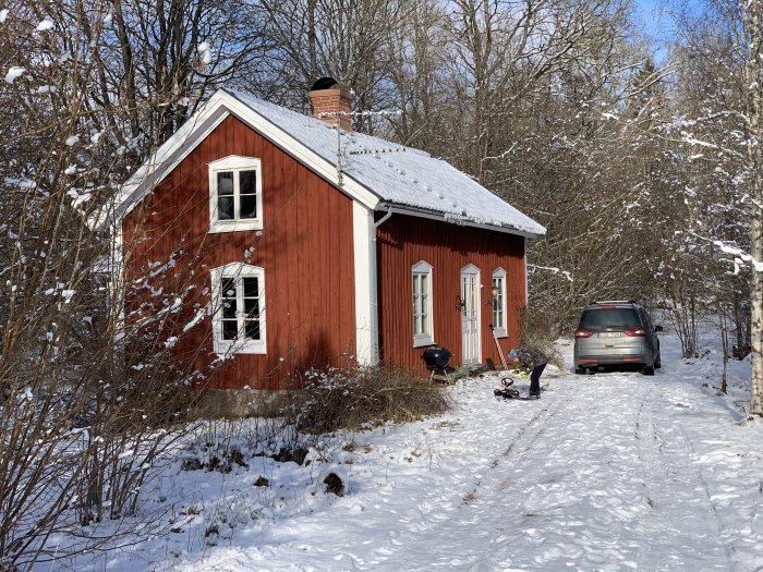 Rött torp med vita knutar i snöig skogsmiljö, barn och bil framför huset.