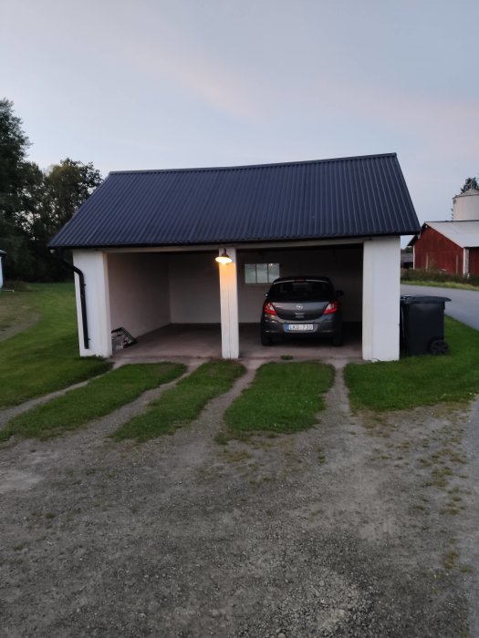 Gammalt garage med öppen carport på grusuppfart, lekablock, plåttak och parkerad bil inne.
