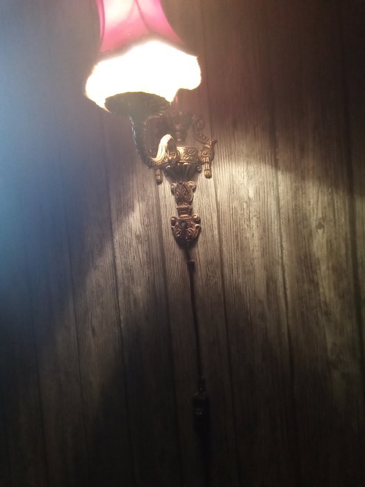 Gammal vägglampa med ornament och röd lampskärm mot en mörk trävägg.