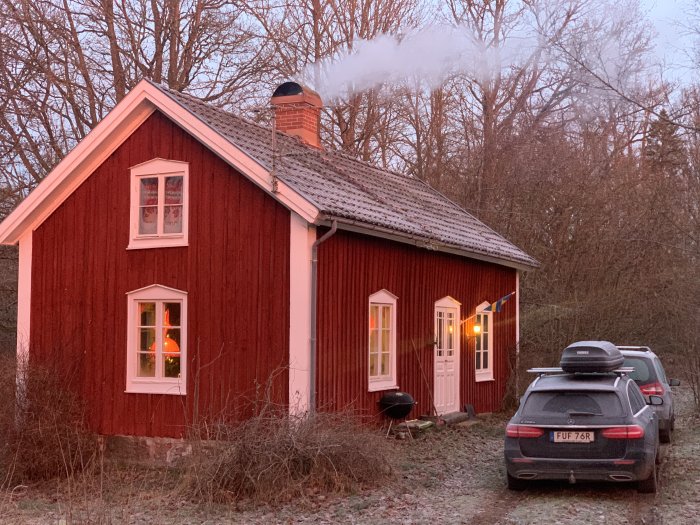 Rödmålat torp med vita knutar och tända fönster, bil parkerad framför, vid skymning.