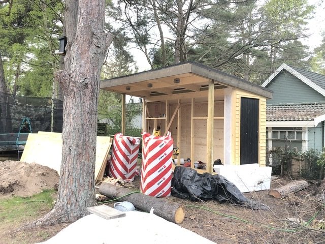 Nybyggd träkonstruktion med tak och panel, placerad i en trädgård, med byggmaterial runtomkring.