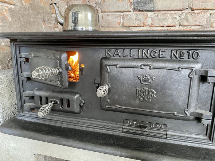 Klassisk gjutjärnsspis märkt "KALLINGE Nº 10" med öppen lucka visar brinnande ved, en tekanna står ovanpå.