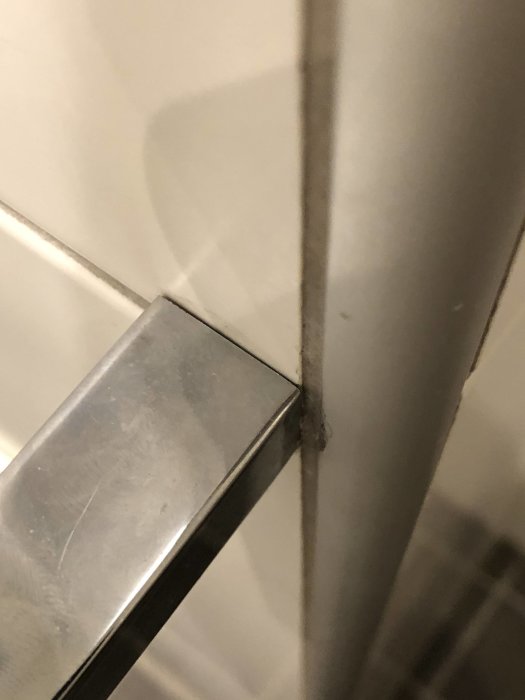 Metallisk pappershållare fäst vid vit vägg med synlig fogskada och orange missfärgning vid kakelfogen.