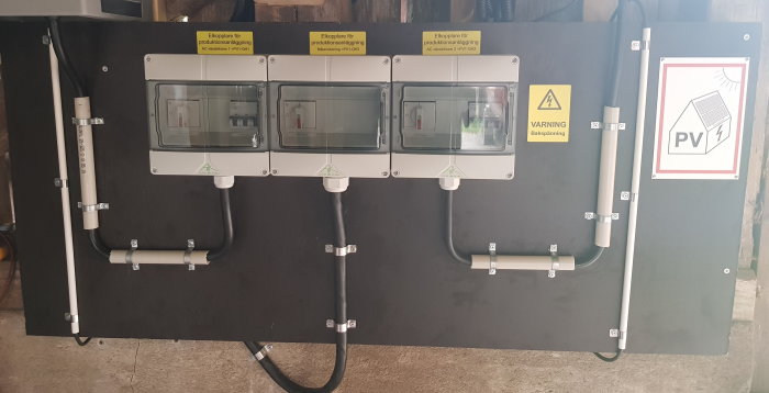 Elektriskt kapslingsskåp med tre elskåp och varningsmärke för bakspänning, installerat på vägg med rörledningar.