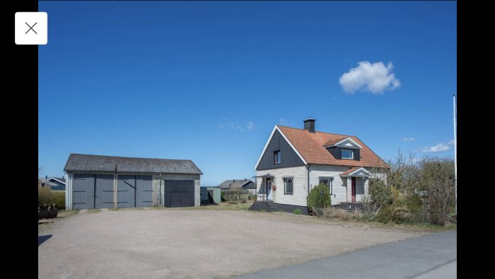 Vit tegelvilla med rött tak, stor garagebyggnad och öppen gård, klar himmel i bakgrunden.