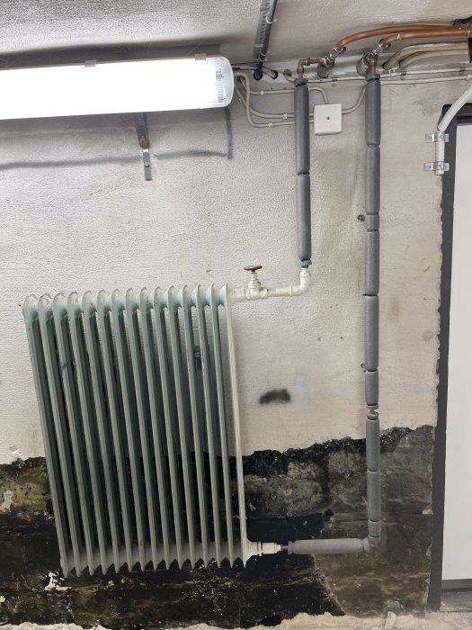 Gammal radiator i ett garage med anslutande rör och T-kopplingar mot en sliten vägg.